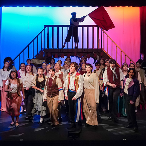 A photo from our 2025 teen summer production of Les Miserables. It shows nearly the full cast of 40 teens singing out to the audience at the end of act one with a large red flag waving behind them all.