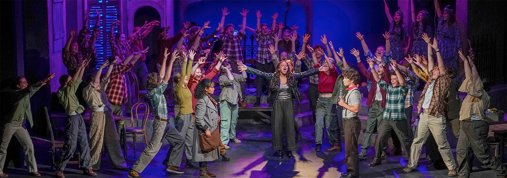Dear Evan Hansen A photo of the cast of Dear Evan Hansen standing in a line across the stage, with Evan Hansen in the middle, closer to the audience. He is in a spotlight with everyone else around hime in shadows. Behind them on the walls is projected a video of swaying tree branches, like a person is looking up at them laying on the ground.
